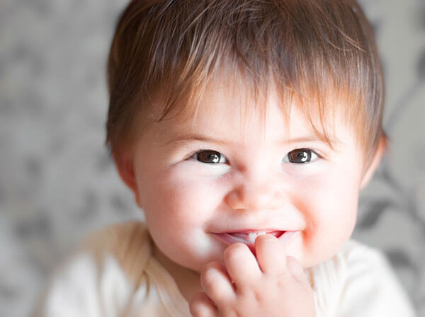 Advizi - La photo représente un bébé souriant avec ses doigt vers la bouche. On distingue ses petites dents. Les yeux sont très expressifs. La photo est prise en gros plan.