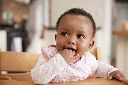 Advizi - La photo représente un bébé fille qui se mange les doigts. Elle est assise sur chaise haute.