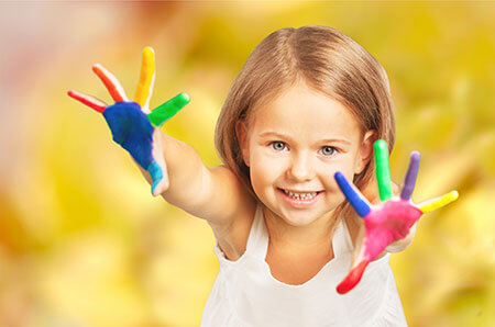 Advizi - La photo représente une petite fille d'environ 4 ans les mains en l'air qui sourit. Ses mains sont peinturées multicolores.